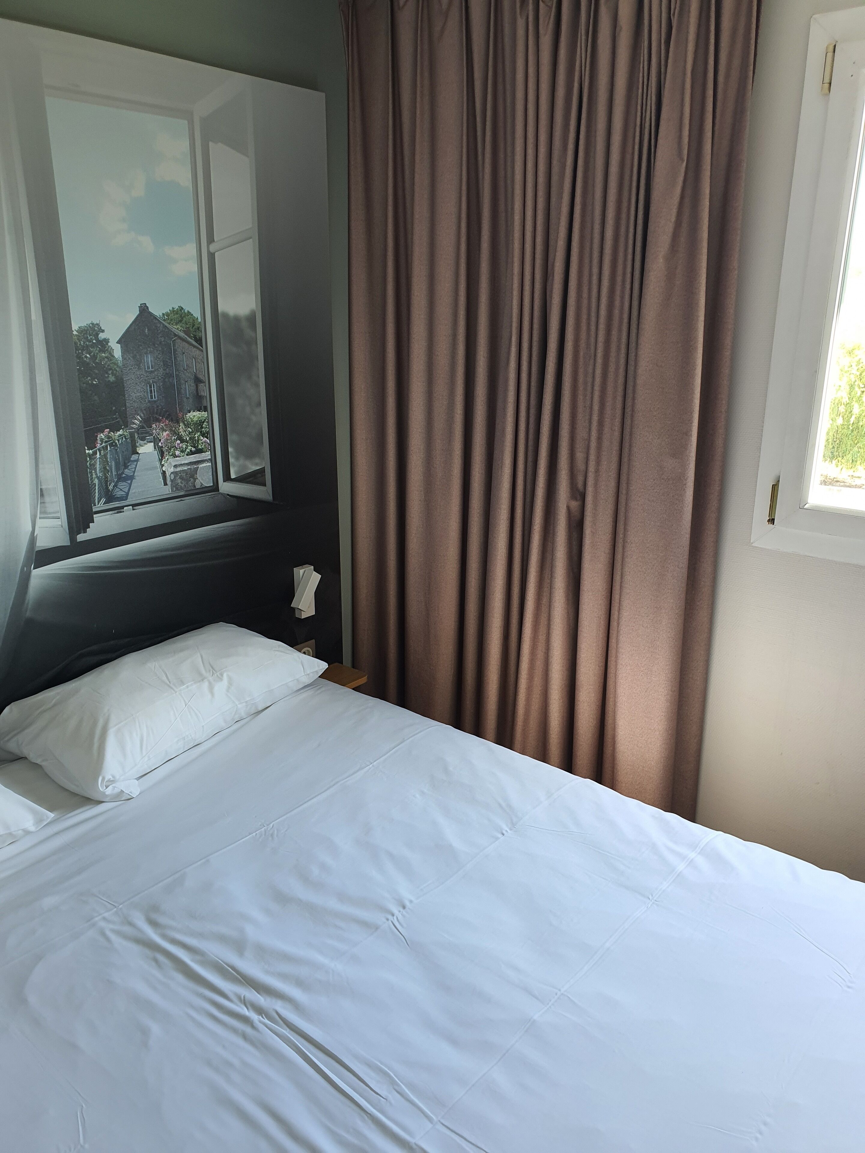 Un lit avec du linge de lit blanc, une fenêtre donnant sur un bâtiment en pierre et des rideaux marron.