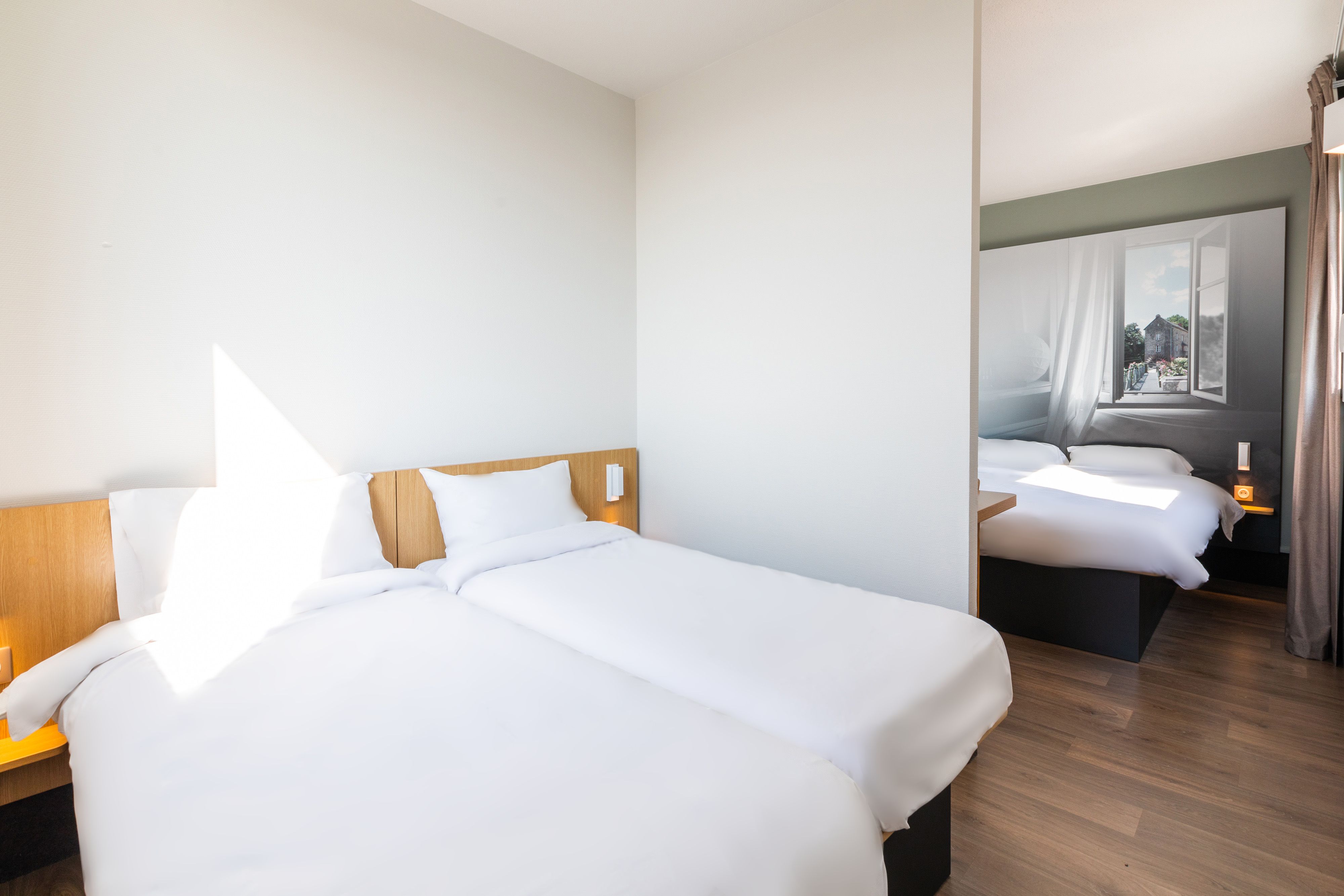 A hotel room with two beds, a wooden headboard, a nightstand, and a window with curtains.