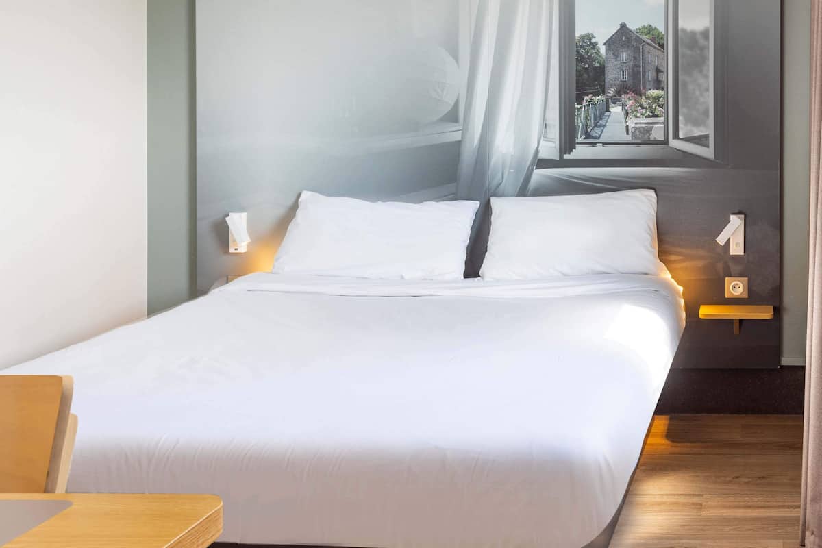 A neatly made bed with white linens, a bedside table with a lamp, and a view of a building through sheer curtains.