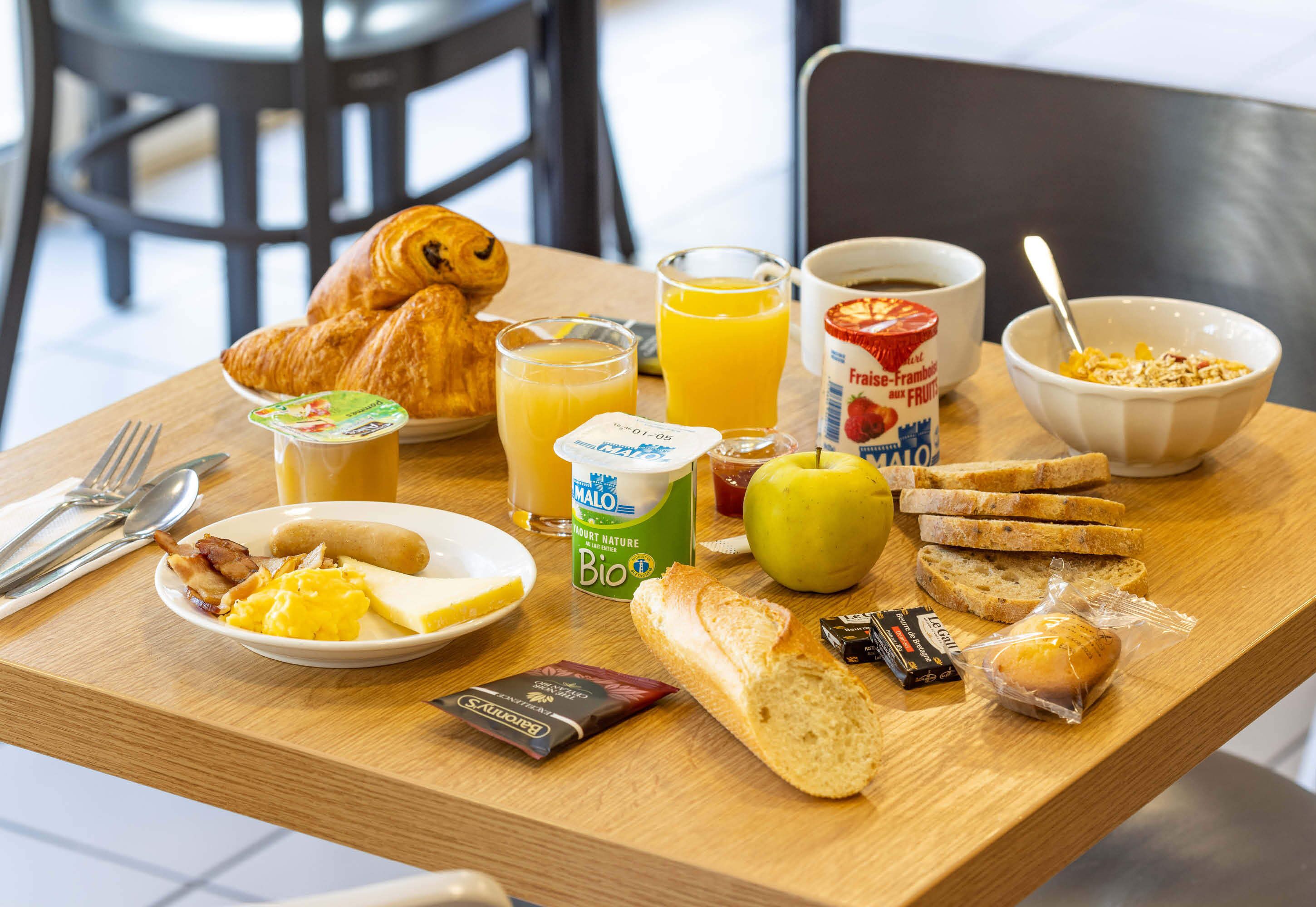 Una mesa de desayuno con croissants, jugo de naranja, una taza de café, un tazón de cereal, un plato con huevos, tostadas, un sándwich y una botella de leche.