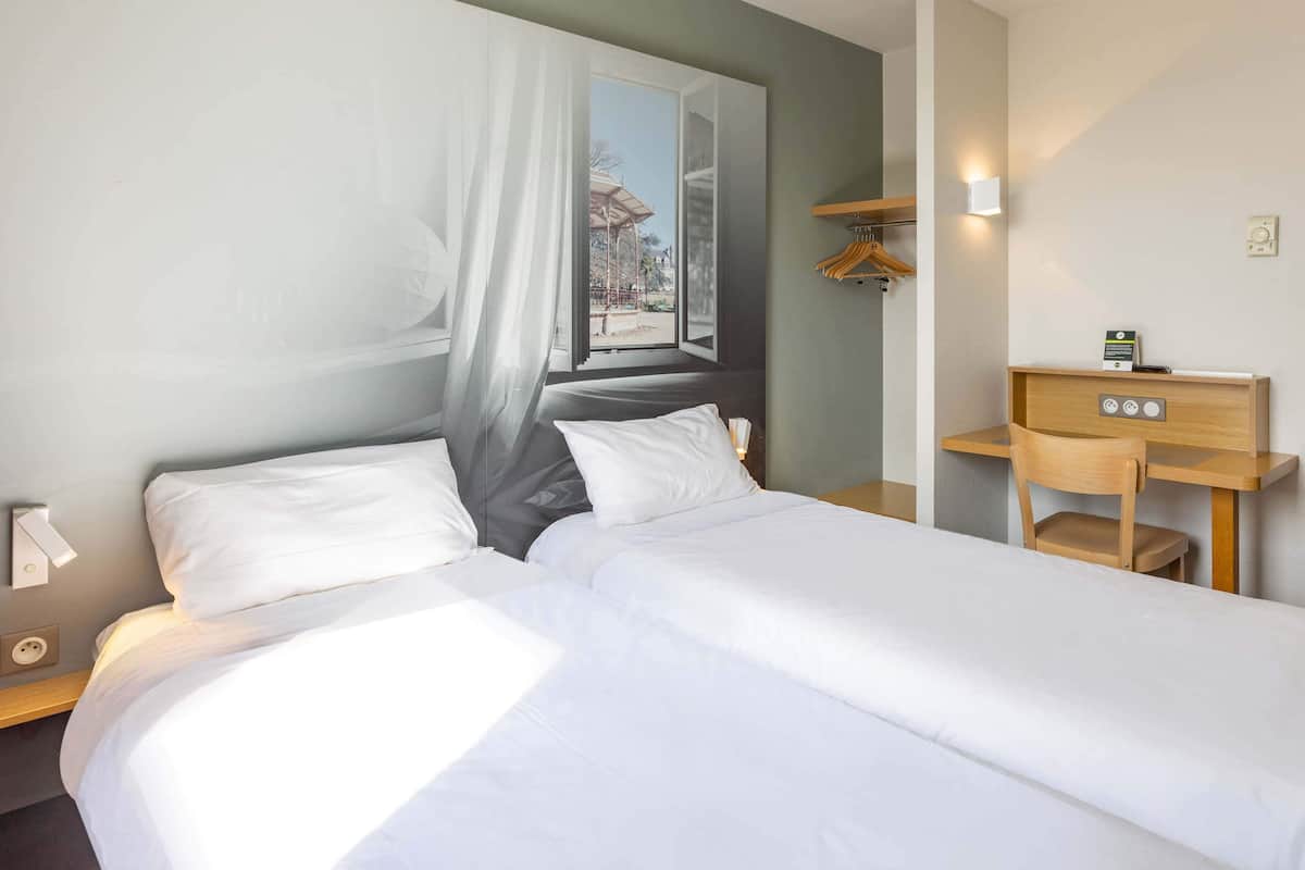 A hotel room with a bed, a desk, and a chair. There is a window with a view of a building and a tree.