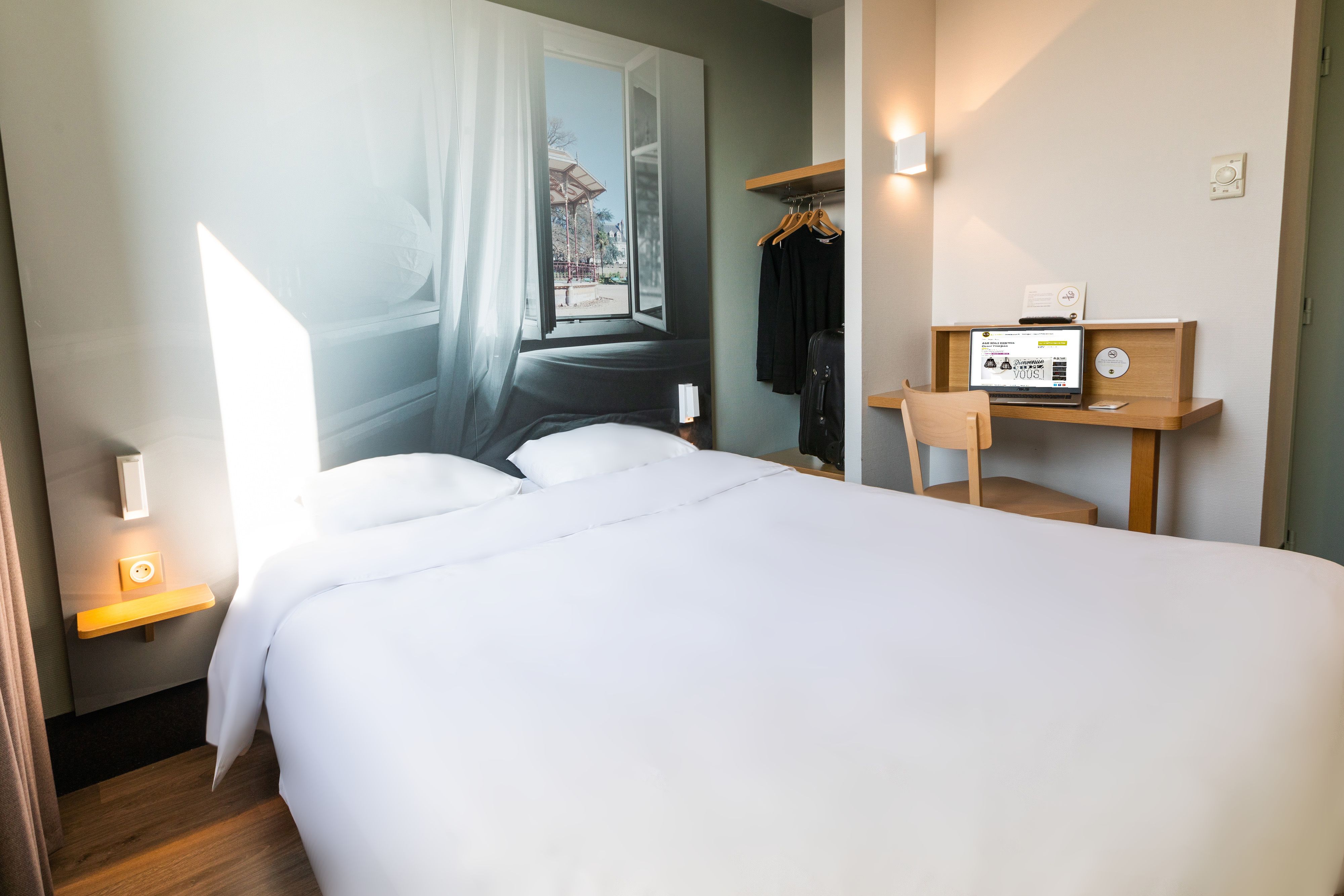 Hotellihuone, jossa on sänky, työpöytä tietokoneella, naulakko ja ikkuna, jossa on verhot.