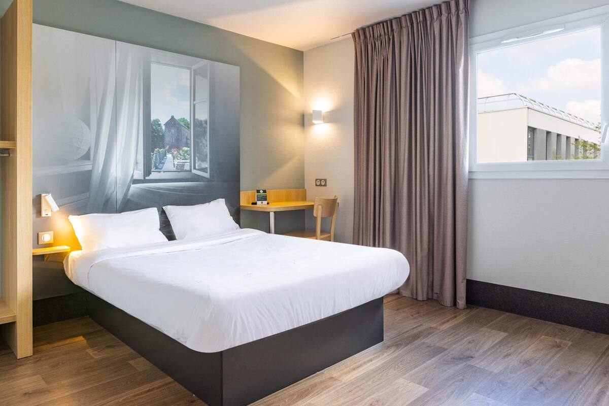 A modern hotel room with a large bed, a desk, a chair, and a view of a building through the window.