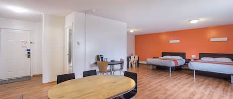A hotel room with a wooden dining table, black chairs, a bed with a red headboard, and a small desk with a chair.
