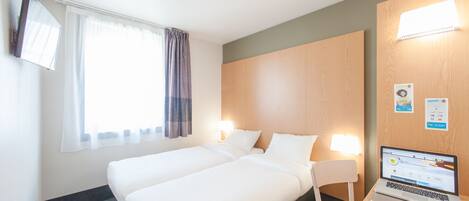 A hotel room with two beds, a desk with a laptop, a chair, a window with curtains, and a wall-mounted TV.