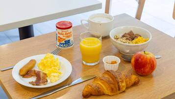 Una mesa de desayuno con huevos revueltos, tocino, cruasanes, un vaso de jugo de naranja, un tazón de cereal, un envase de yogurt y una manzana.