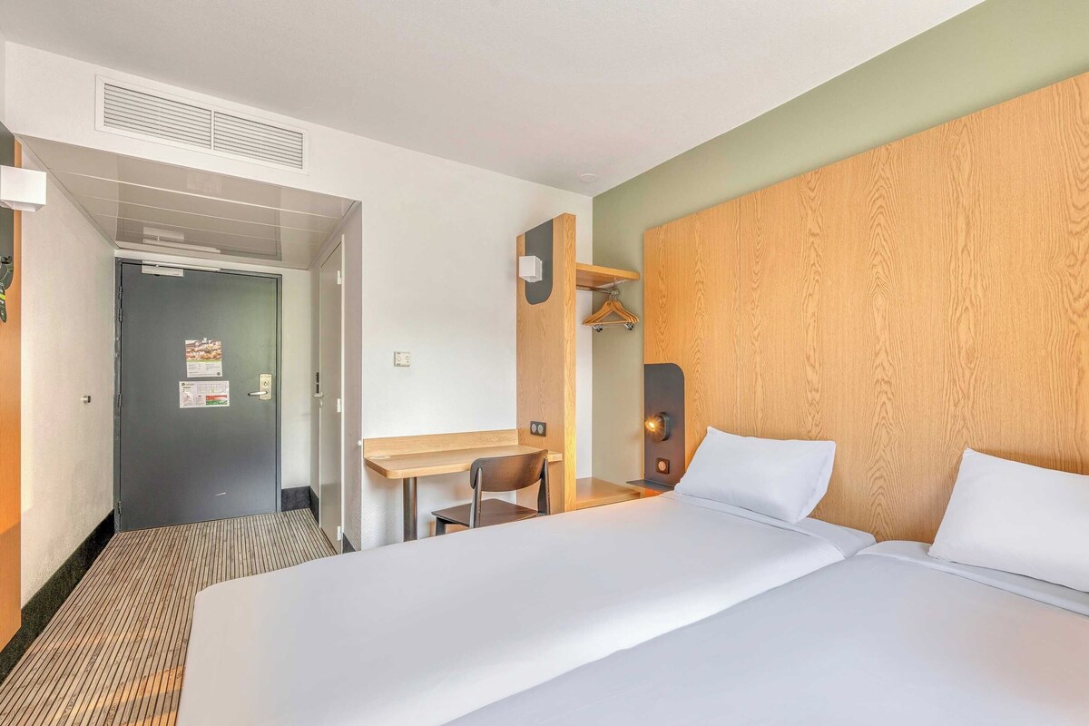 Habitación de hotel con una cama grande, un escritorio y una silla. Se observa una puerta con un letrero, una estantería en la pared y un cabecero de madera.