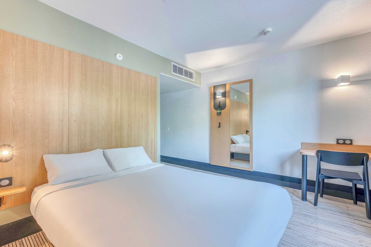 Une chambre d’hôtel avec un grand lit, un bureau, une chaise, un miroir et un luminaire fixé au mur.