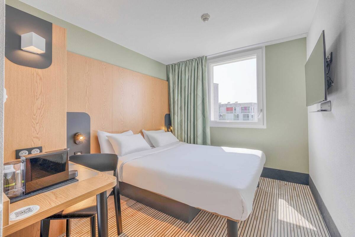 Une chambre d’hôtel équipée d’un lit, d’un bureau avec un ordinateur, d’une fenêtre avec des rideaux et d’un téléviseur à écran plat fixé au mur.