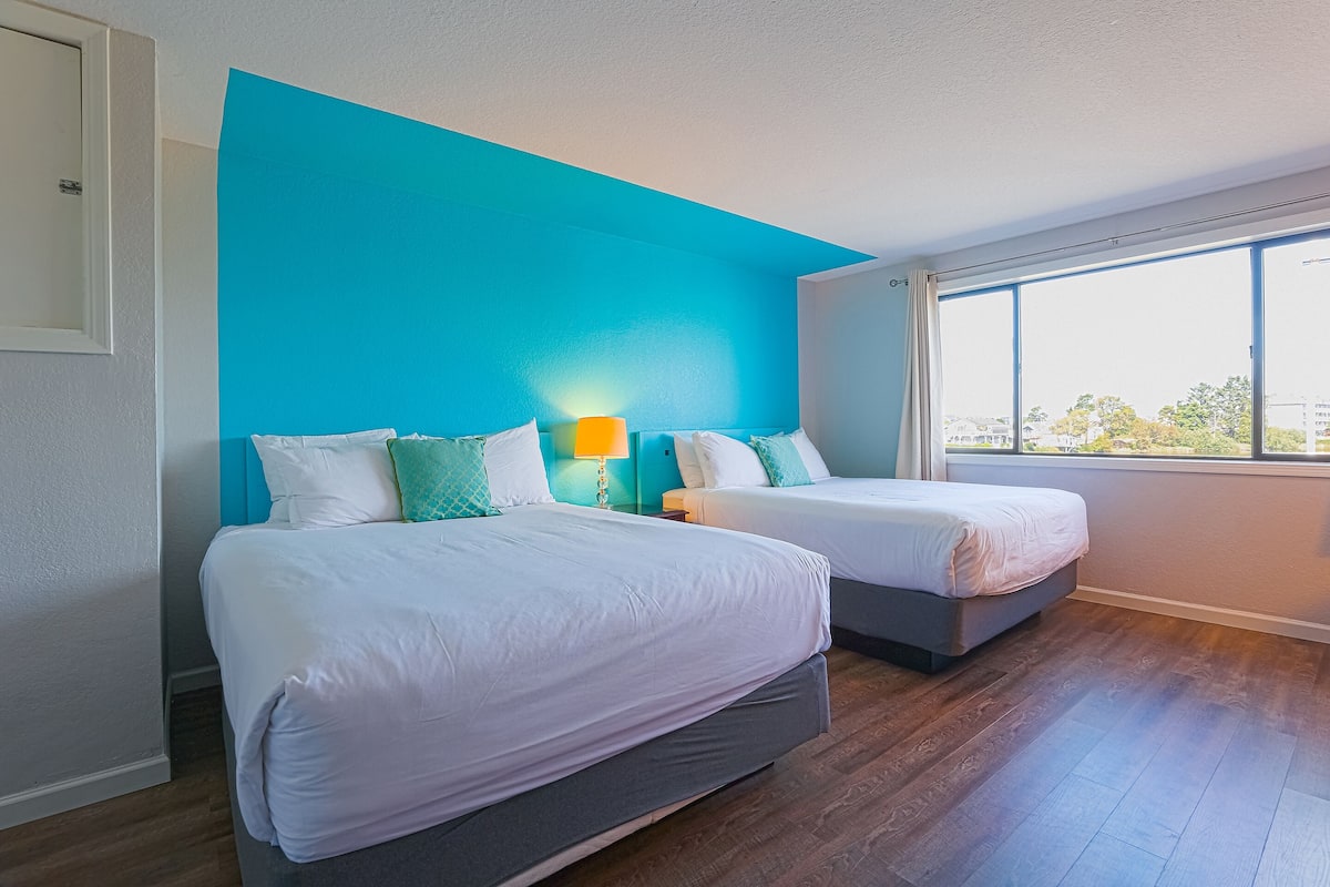 A hotel room with two beds, a teal accent wall, a large window, and a wooden floor.