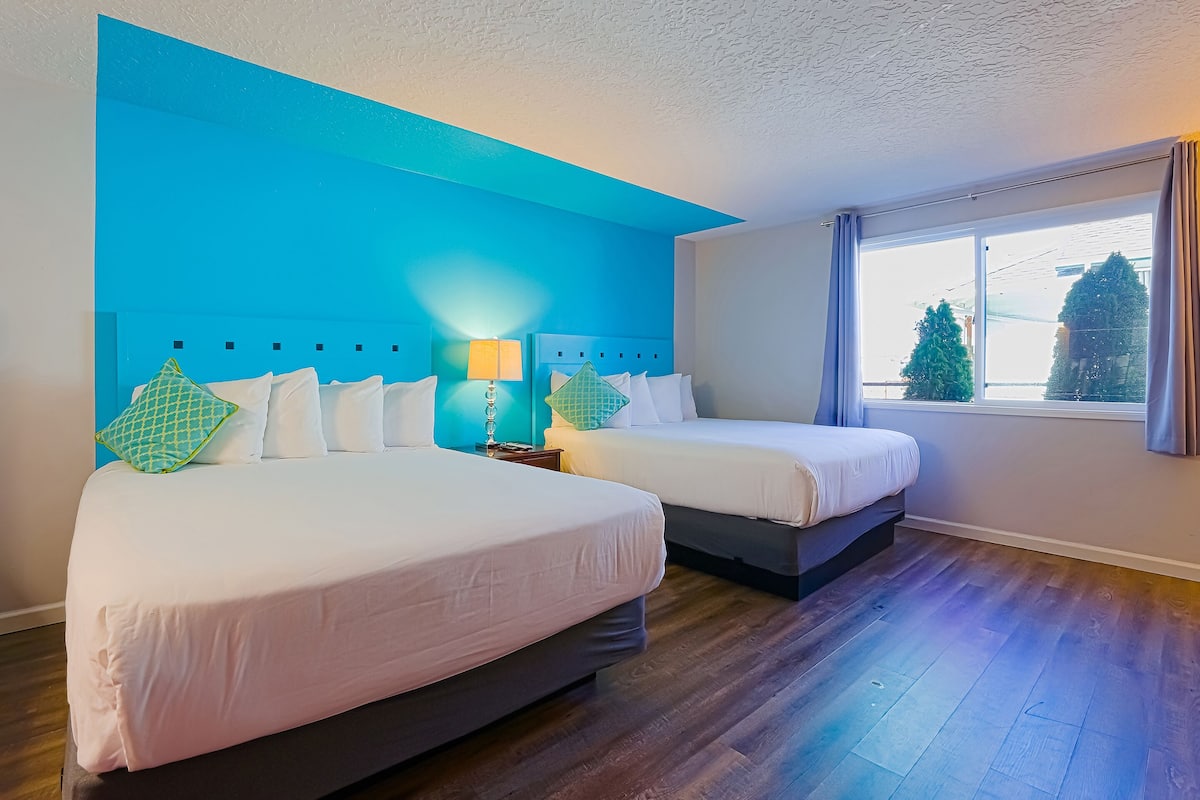 A hotel room with two beds, a large window with curtains, and a view of trees outside.
