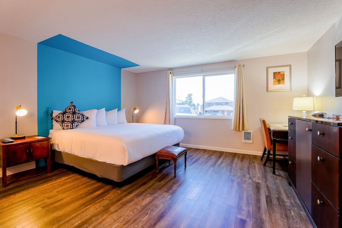 A hotel room with a bed, bedside table, lamp, window, desk, chair, and a dresser.
