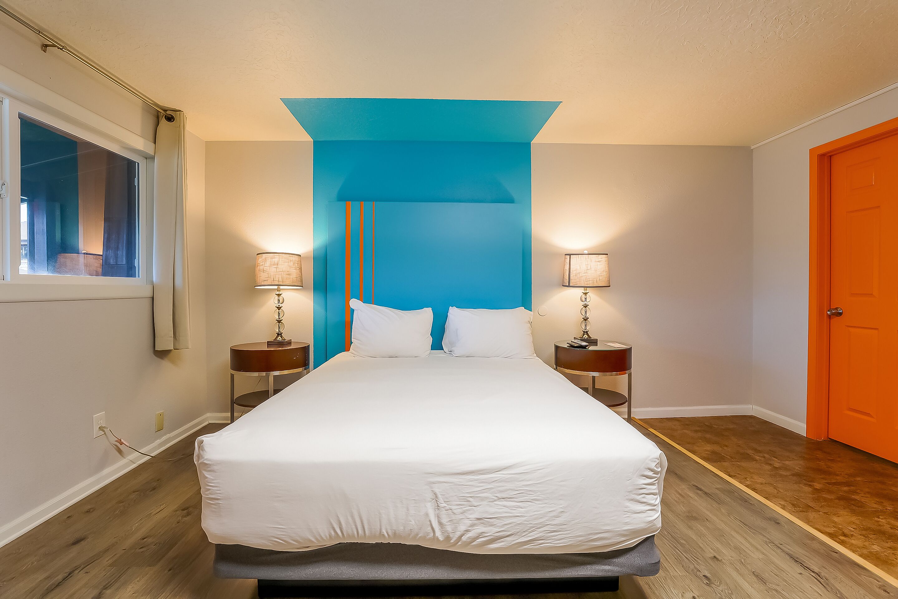 A bedroom with a large bed, two bedside tables with lamps, a window with curtains, and an orange door.