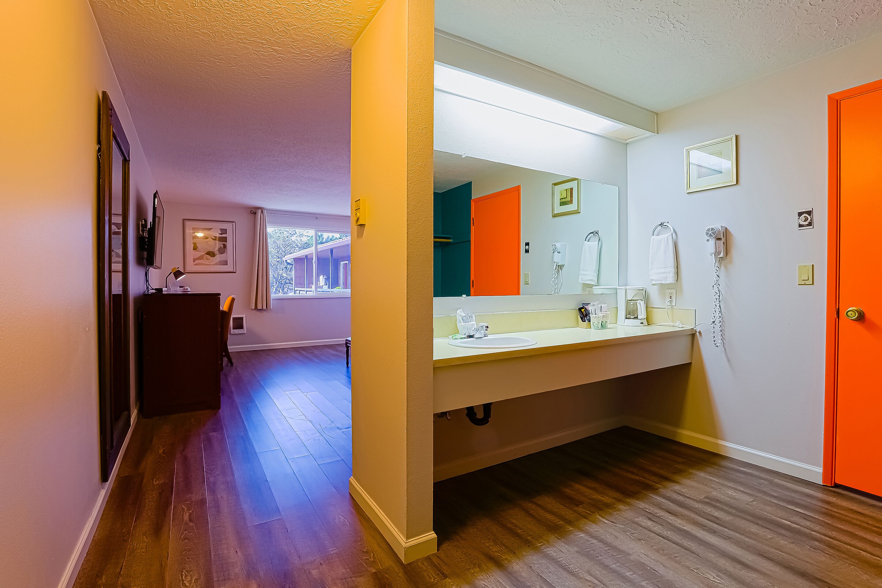 A bathroom with a large mirror, a sink, and a toilet. There is a door with an orange frame and a towel rack with a towel.