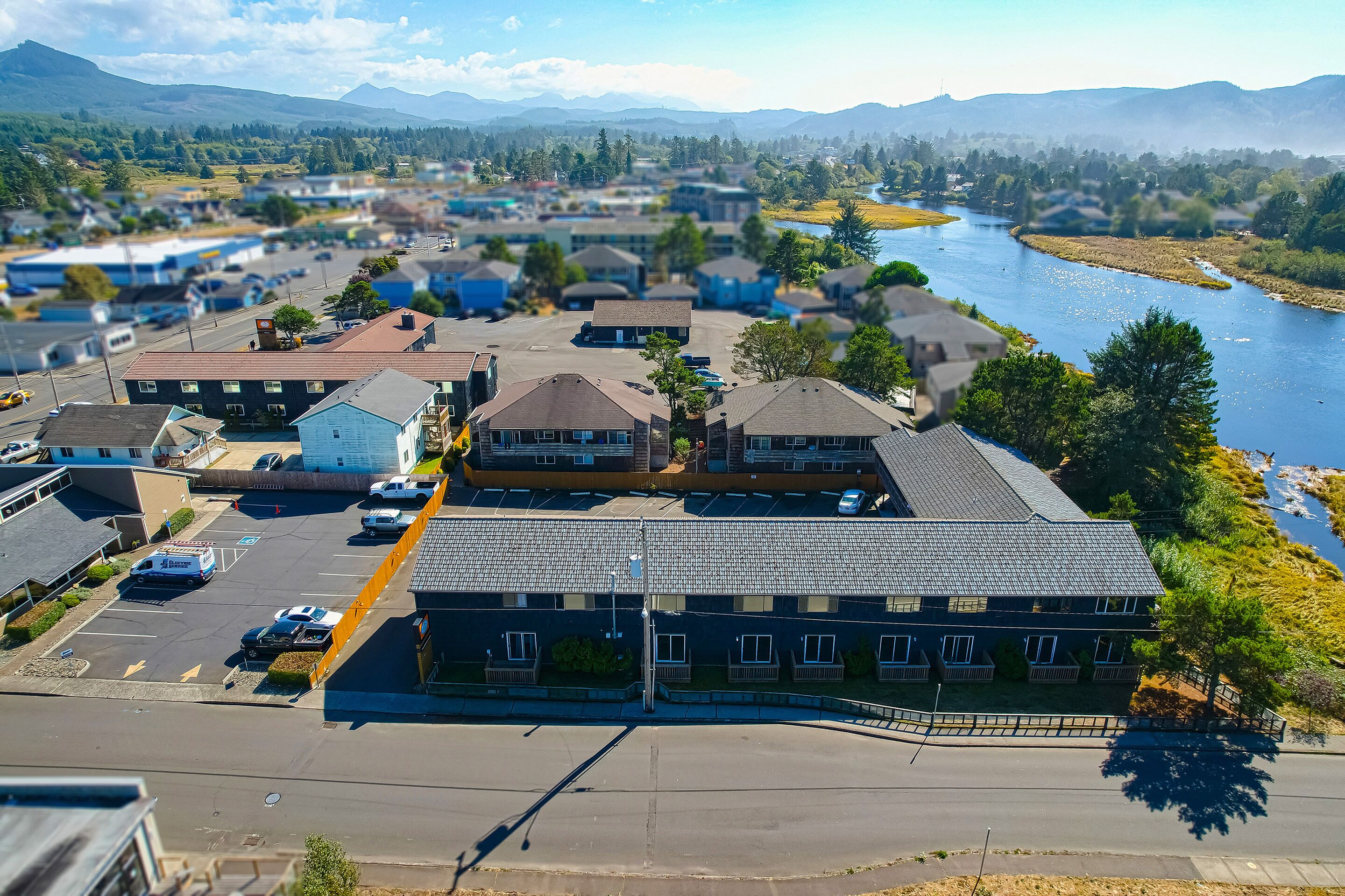 A multi-story hotel with a parking lot, situated near a river with mountains in the background.