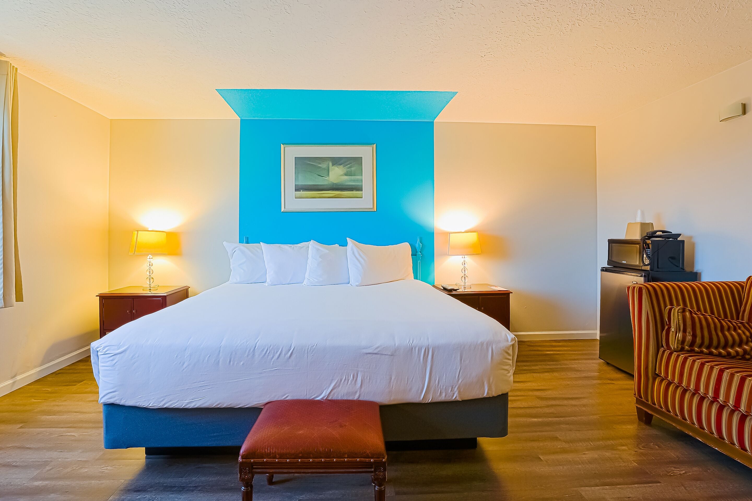 A hotel room with a large bed, two bedside lamps, a small table with a lamp, a television, and a striped armchair.
