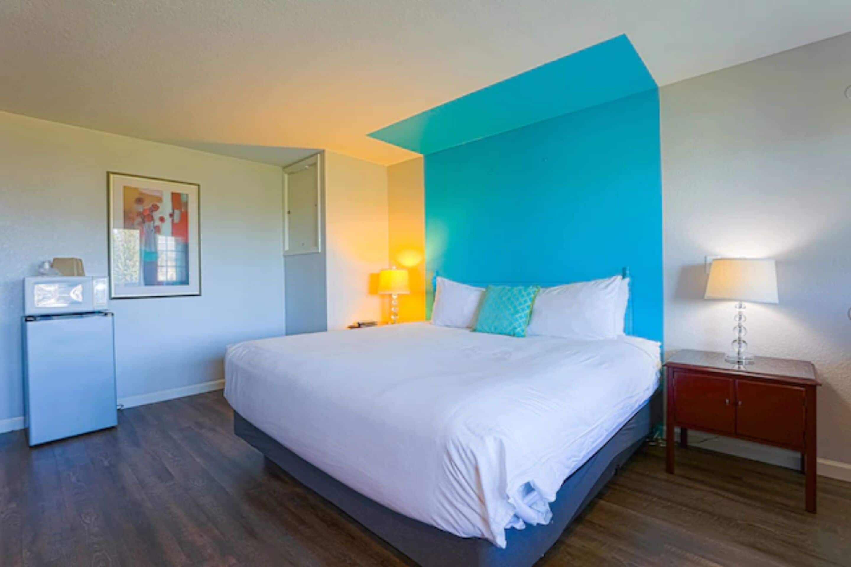 A hotel room with a large bed, a small desk, a nightstand, a lamp, and a refrigerator.