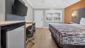 Habitación de hotel con cama, escritorio, silla, televisor y ventana con persianas.