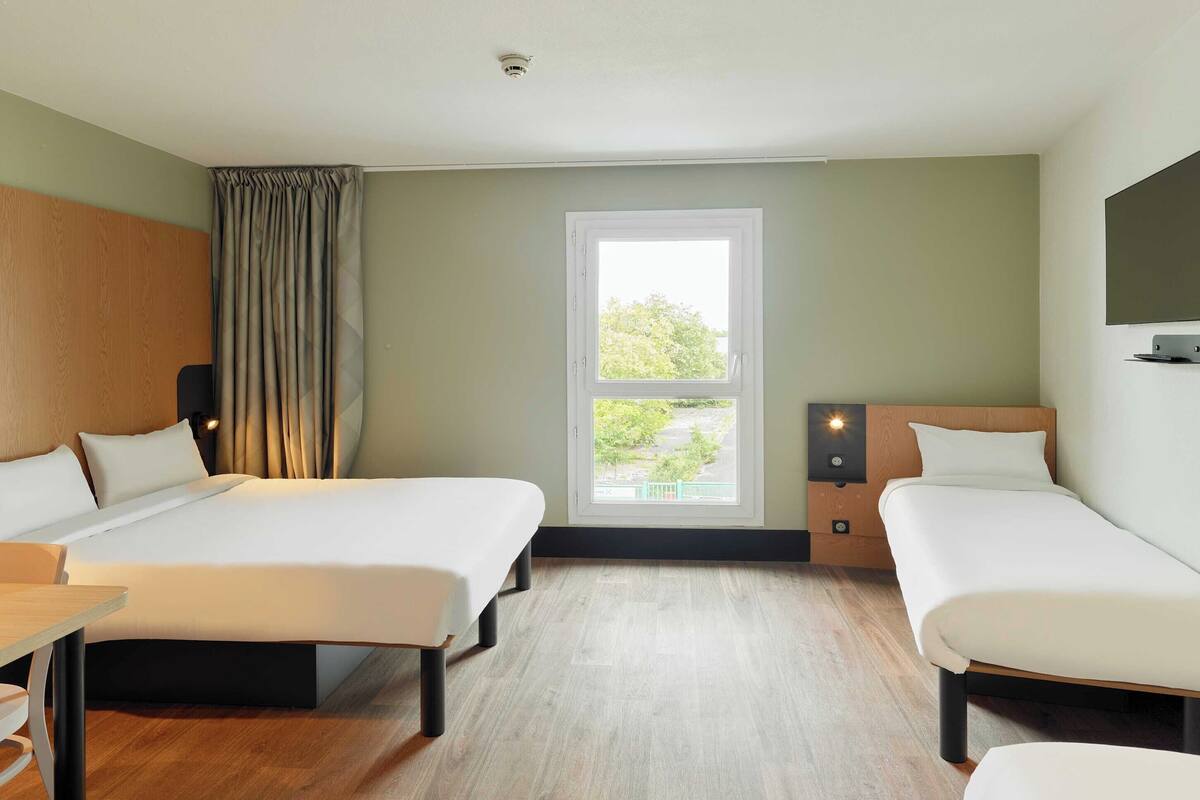 A hotel room with two beds, a desk, a TV, and a window with a view of greenery.