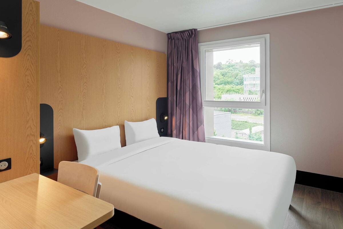 A hotel room with a bed, a wooden headboard, a window with curtains, and a table.