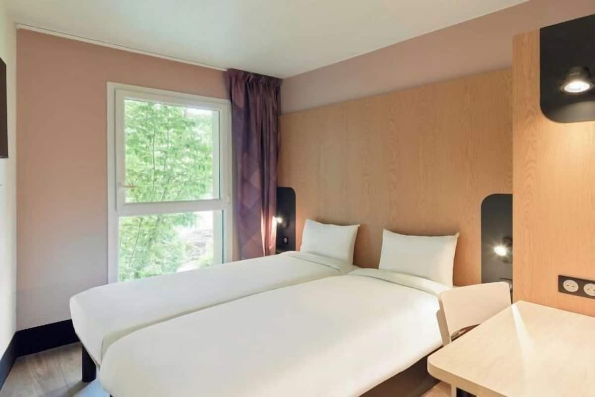 Habitación de hotel con una cama grande, una mesita pequeña, una ventana con cortinas y un aplique de pared.