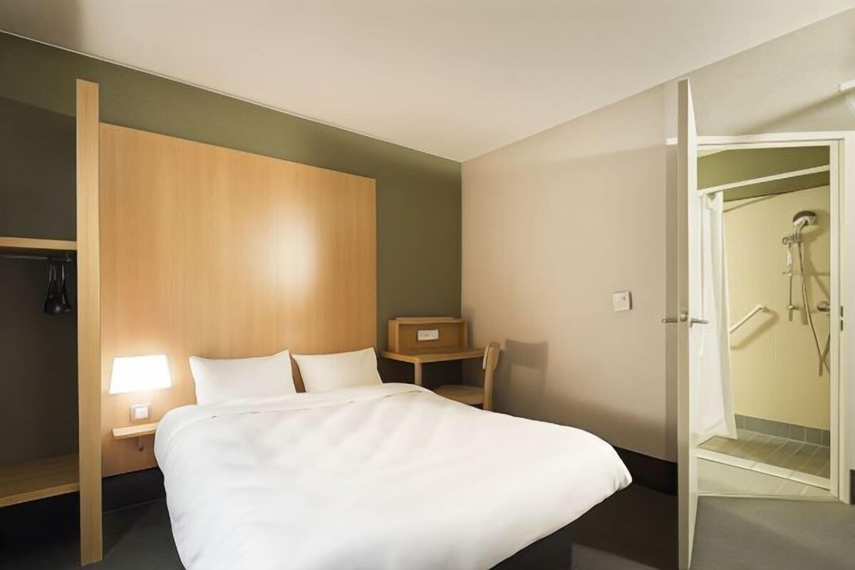 A hotel room with a bed, a nightstand, a wooden wardrobe, a desk, and a shower area.
