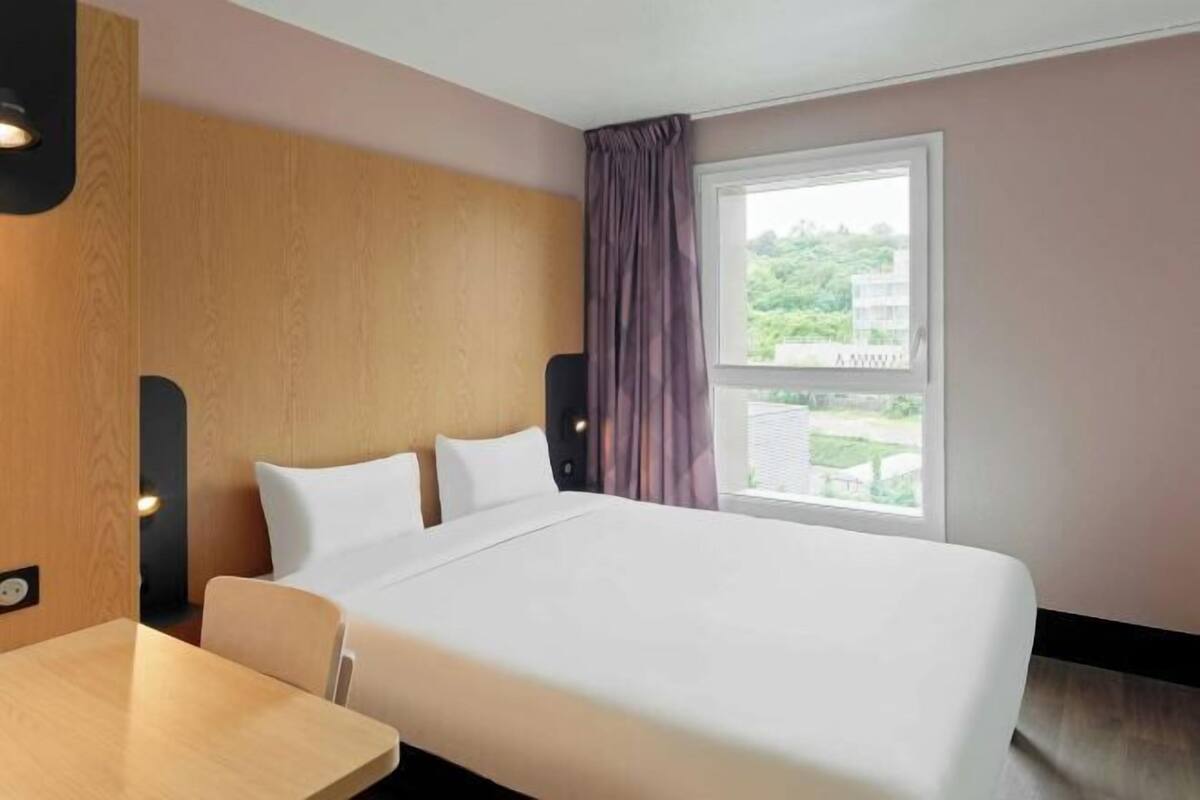 A hotel room with a bed, a table, a window with curtains, and a view of greenery outside.