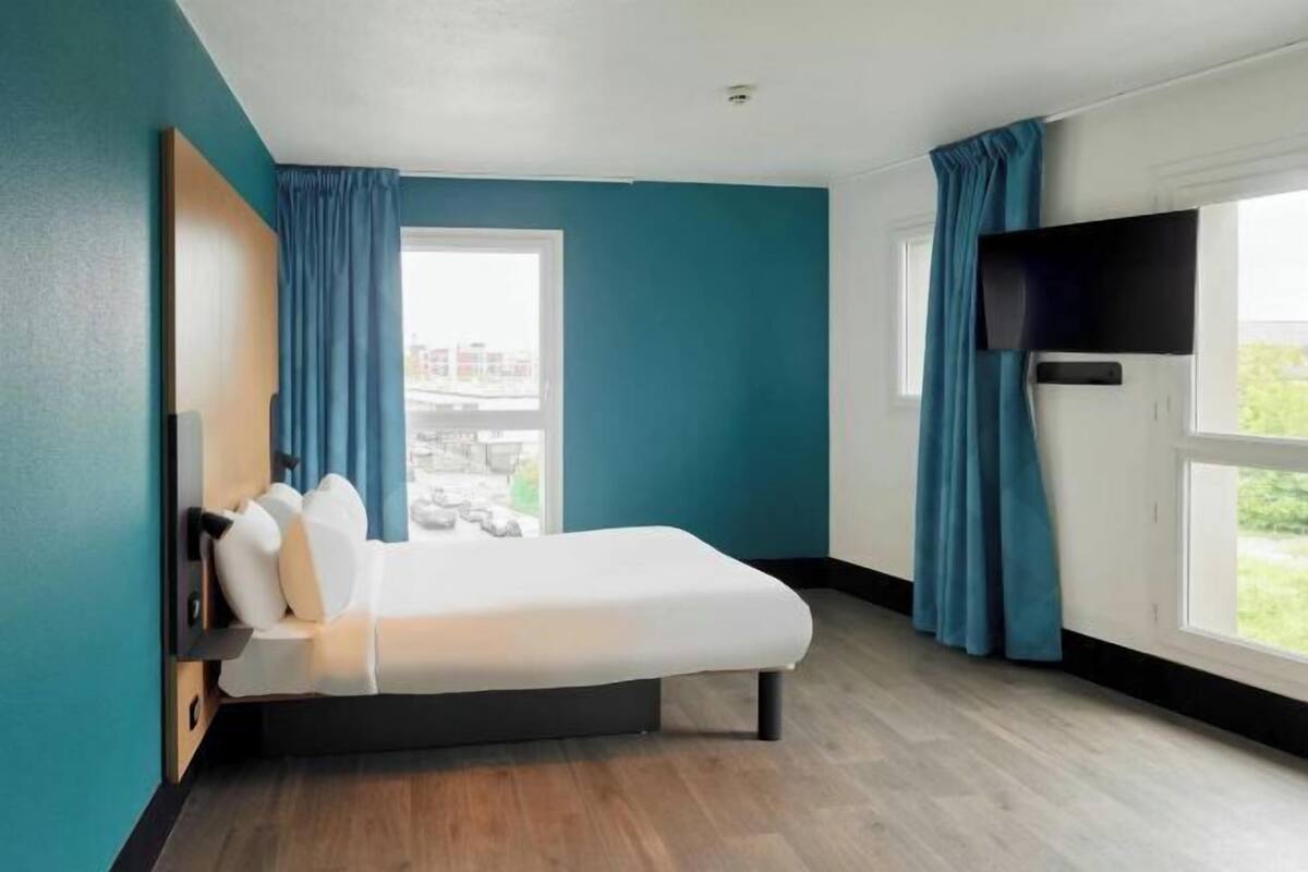 A hotel room with a bed, a TV mounted on the wall, and a window with curtains.