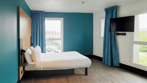 A modern hotel room with a bed, a flat-screen TV, a window with curtains, and a view of the cityscape.