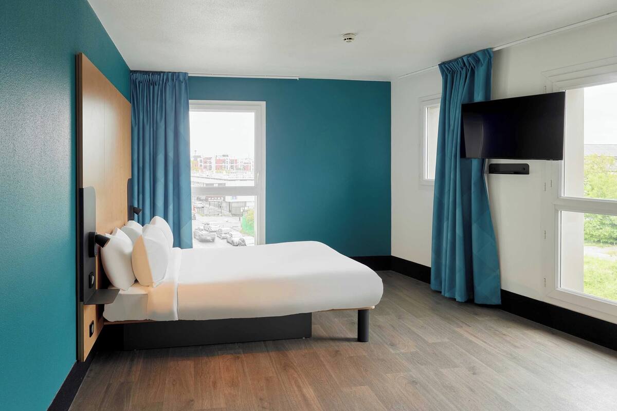 A modern hotel room with a bed, a flat-screen TV, a window with curtains, and a view of the cityscape.