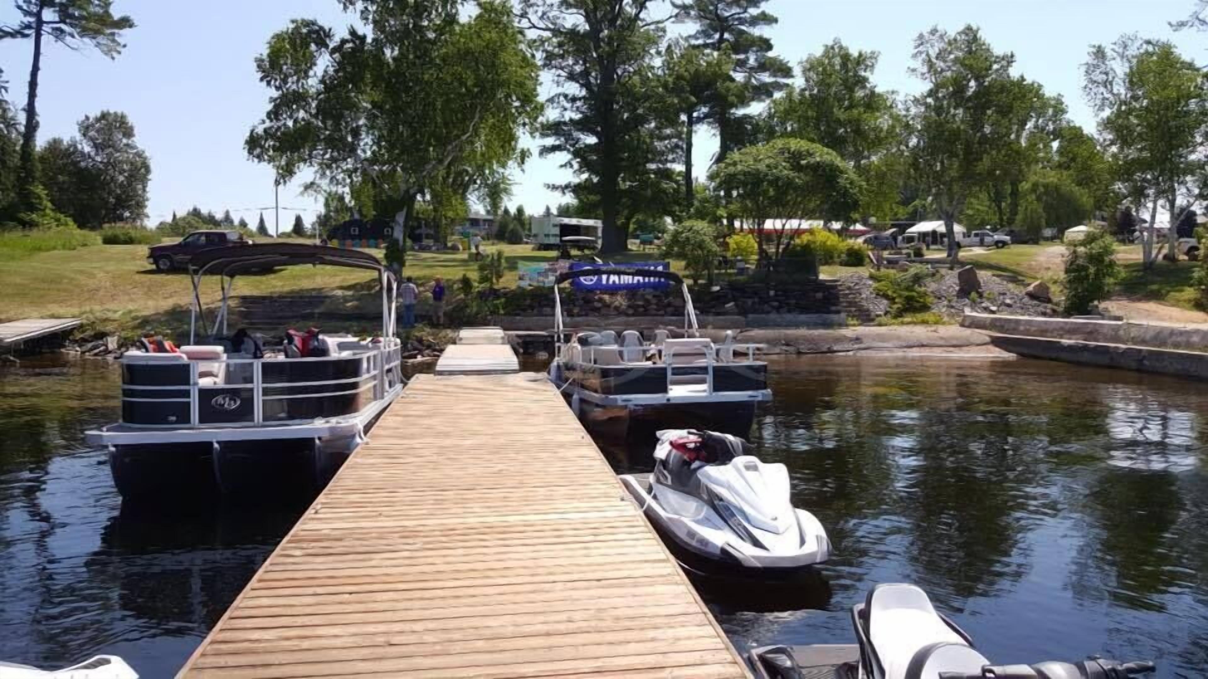 A wooden dock with boats docked at a calm water body, surrounded by greenery and a clear sky.