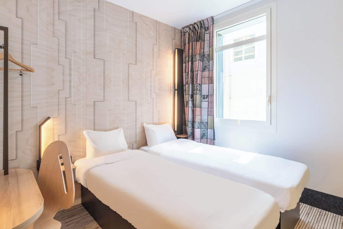 A hotel room with two beds, a wooden chair, a desk, and a patterned curtain.