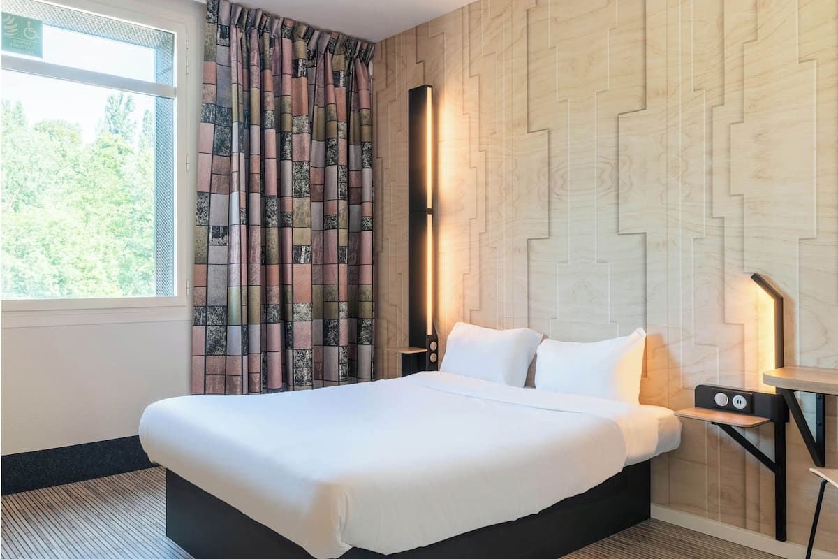 A hotel room with a large bed, a nightstand with a lamp, a window with curtains, and a wall with a geometric pattern.