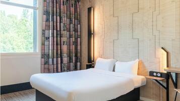 A hotel room with a large bed, a nightstand with a lamp, a window with curtains, and a wall with a geometric pattern.