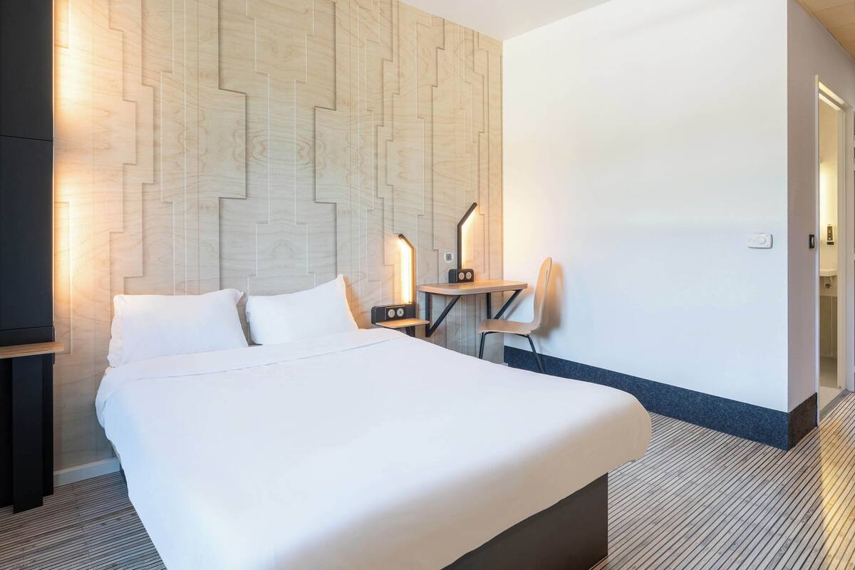 A hotel room with a large bed, a desk with a chair, a wall-mounted lamp, and a wooden accent wall.