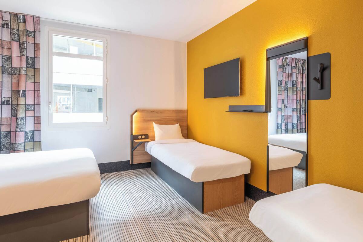 A hotel room with two single beds, a large mirror, a flat-screen TV, and a window with curtains.