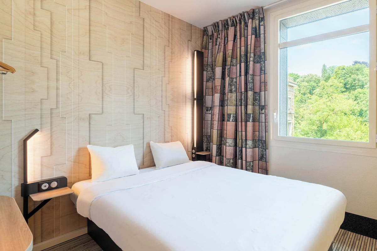 A hotel room with a large bed, a nightstand with a lamp, a window with curtains, and a view of greenery outside.