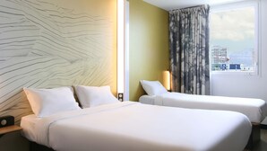 Habitación de hotel con dos camas, un ventanal con vistas a edificios y una montaña, y una pared decorada con una lámpara original.
