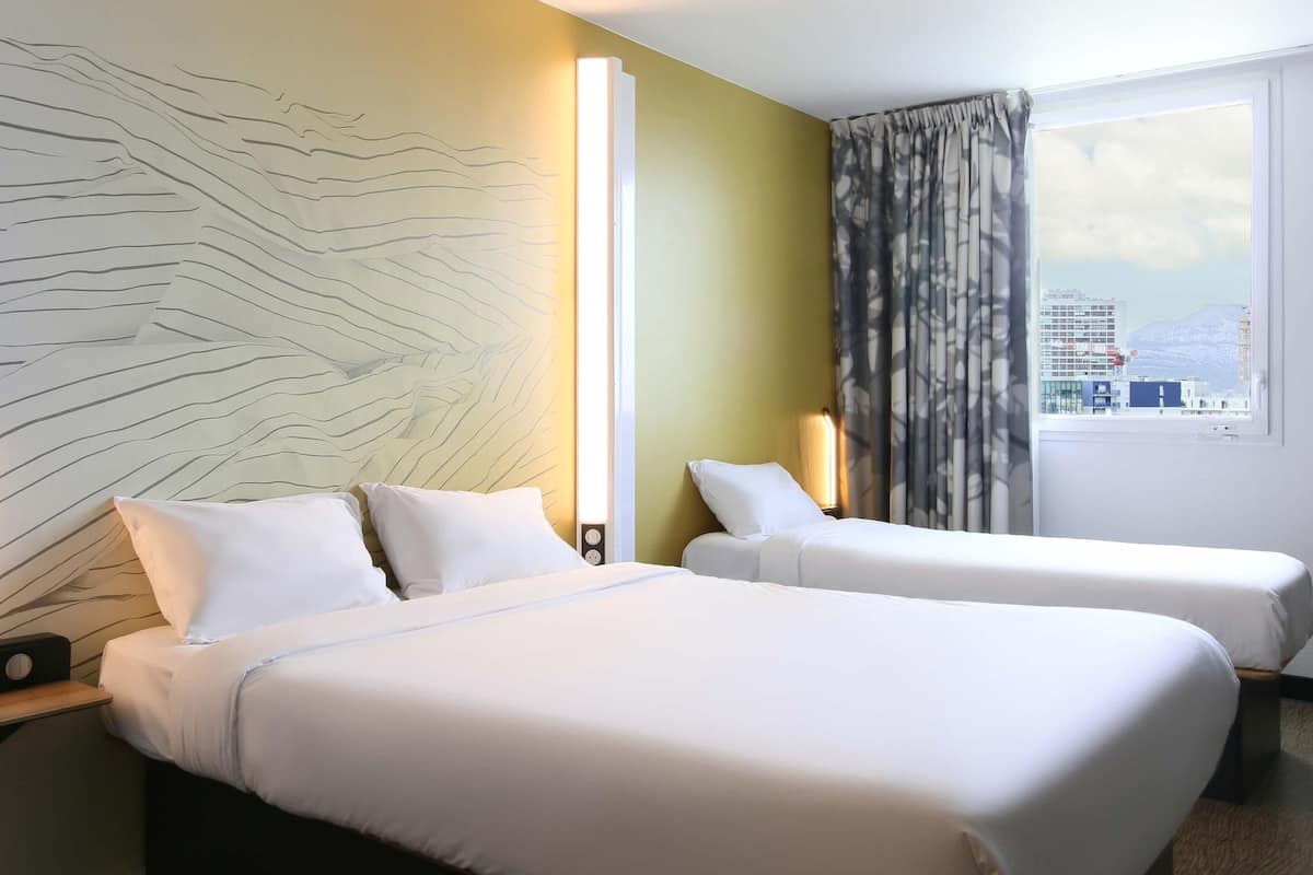 Habitación de hotel con dos camas, un ventanal con vistas a edificios y una montaña, y una pared decorada con una lámpara original.