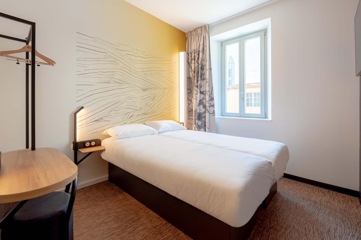 Habitación de hotel con cama, escritorio, silla, cuadro en la pared, ventana con cortinas y vistas al exterior.