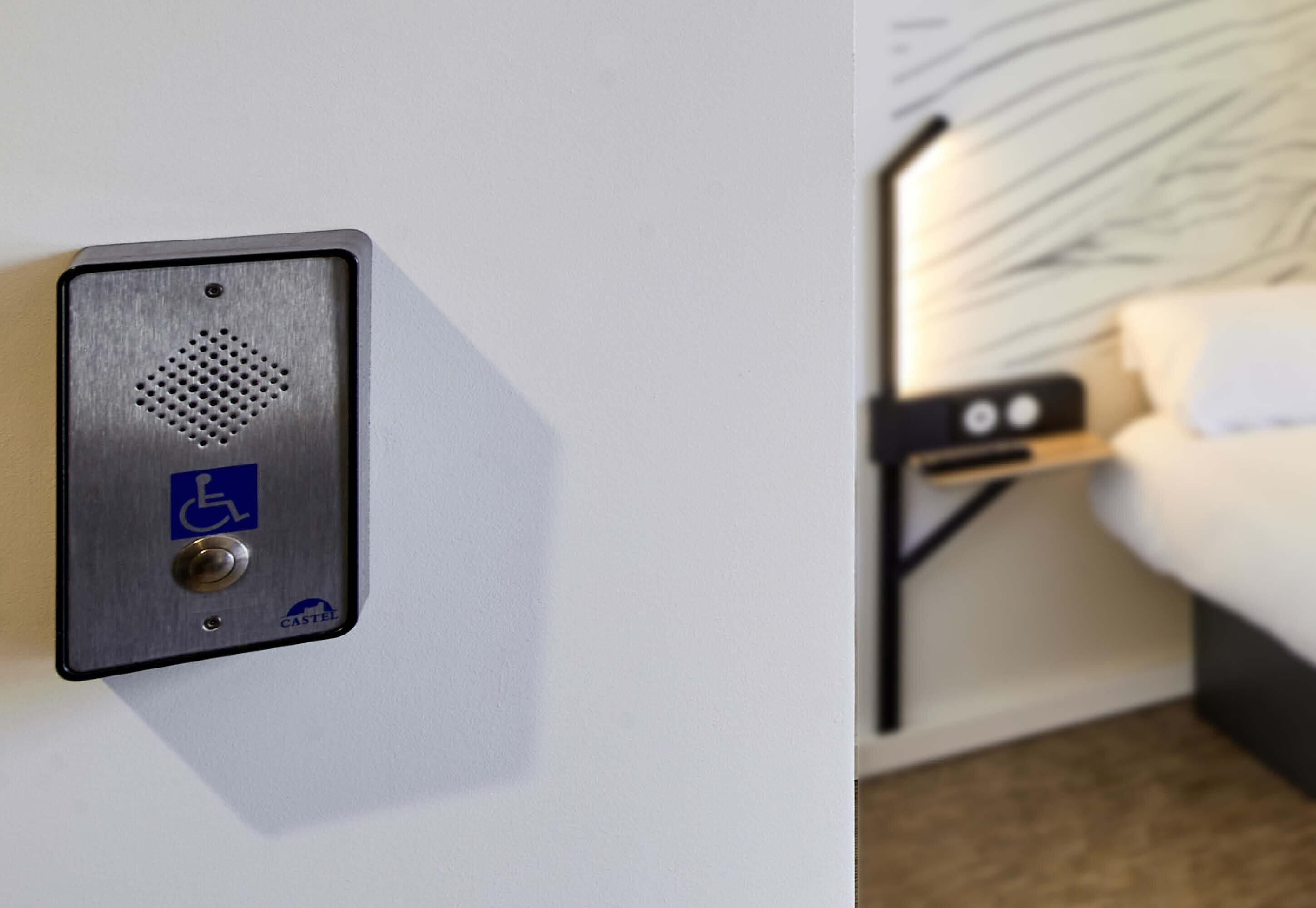 Habitación de hotel con cama, mesita de noche y panel de control en la pared con símbolo de accesibilidad para sillas de ruedas.