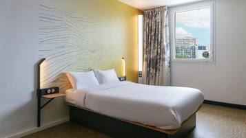 A neatly made bed in a hotel room with a city view, a wall art piece, and a nightstand with a lamp.