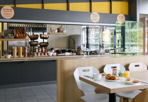 A breakfast area with a counter displaying pastries, bread, and condiments. There are white chairs and a table with breakfast items. - B&B HOTEL Marseille Vélodrome Prado (Marseille)