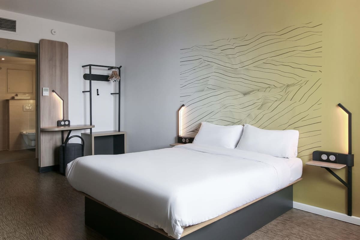 Habitación de hotel moderna con una cama grande, una mesita de noche con lámpara, un estante en la pared y un baño visible a través de una puerta abierta.