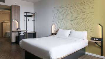 Habitación de hotel moderna con una cama grande, una mesita de noche con lámpara, un estante en la pared y un baño visible a través de una puerta abierta.