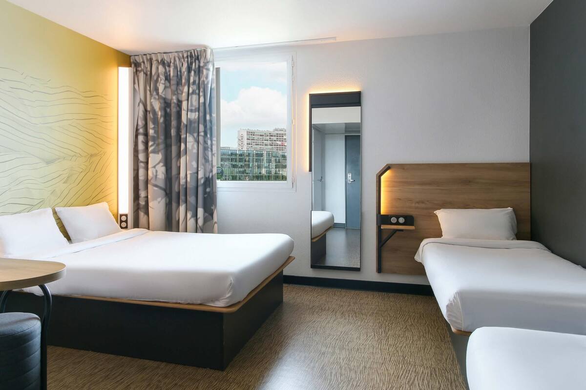 Habitación de hotel con dos camas, una mesita, una silla, un ventanal grande con cortinas y un espejo en la pared.