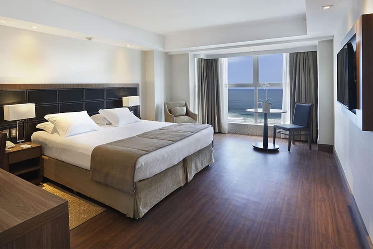 Una habitación de hotel moderna con una cama grande, mesitas de noche con lámparas, un escritorio con una silla, un televisor y un balcón con vista al océano.