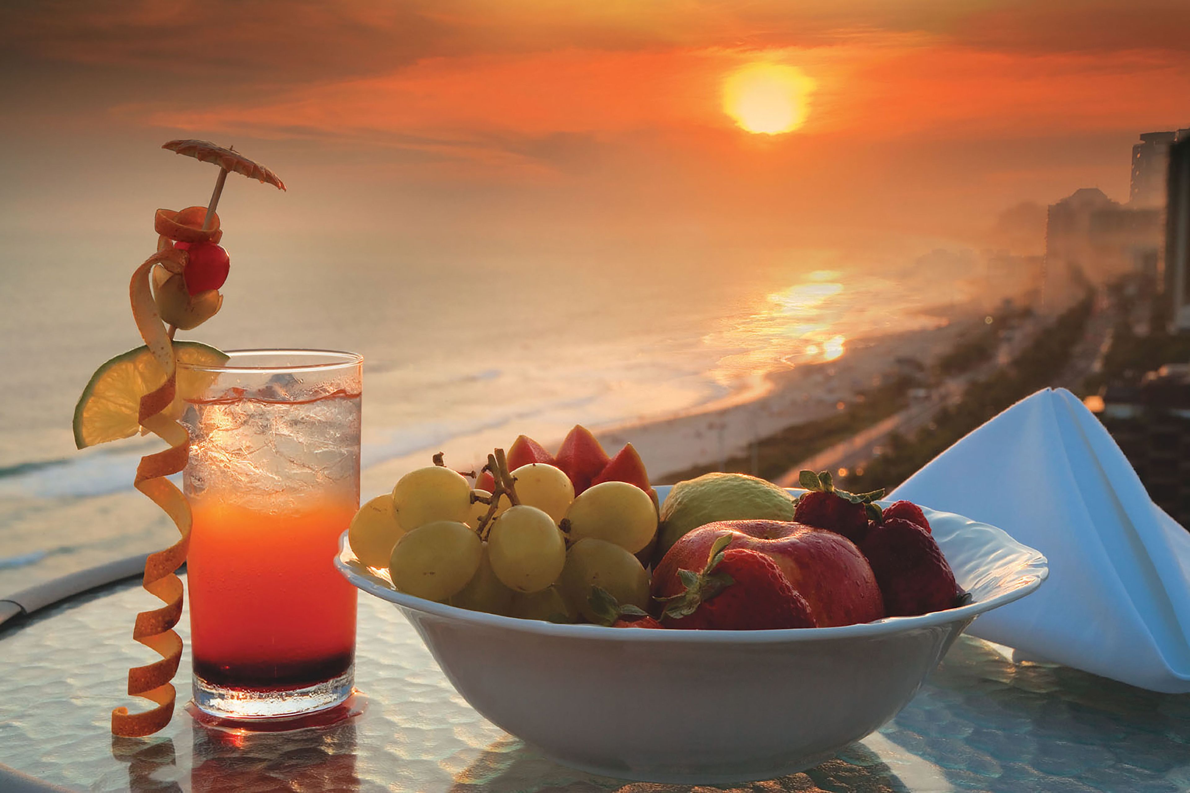 Una vista al atardecer con una copa de bebida roja, un bol de fruta y una servilleta sobre una mesa.
