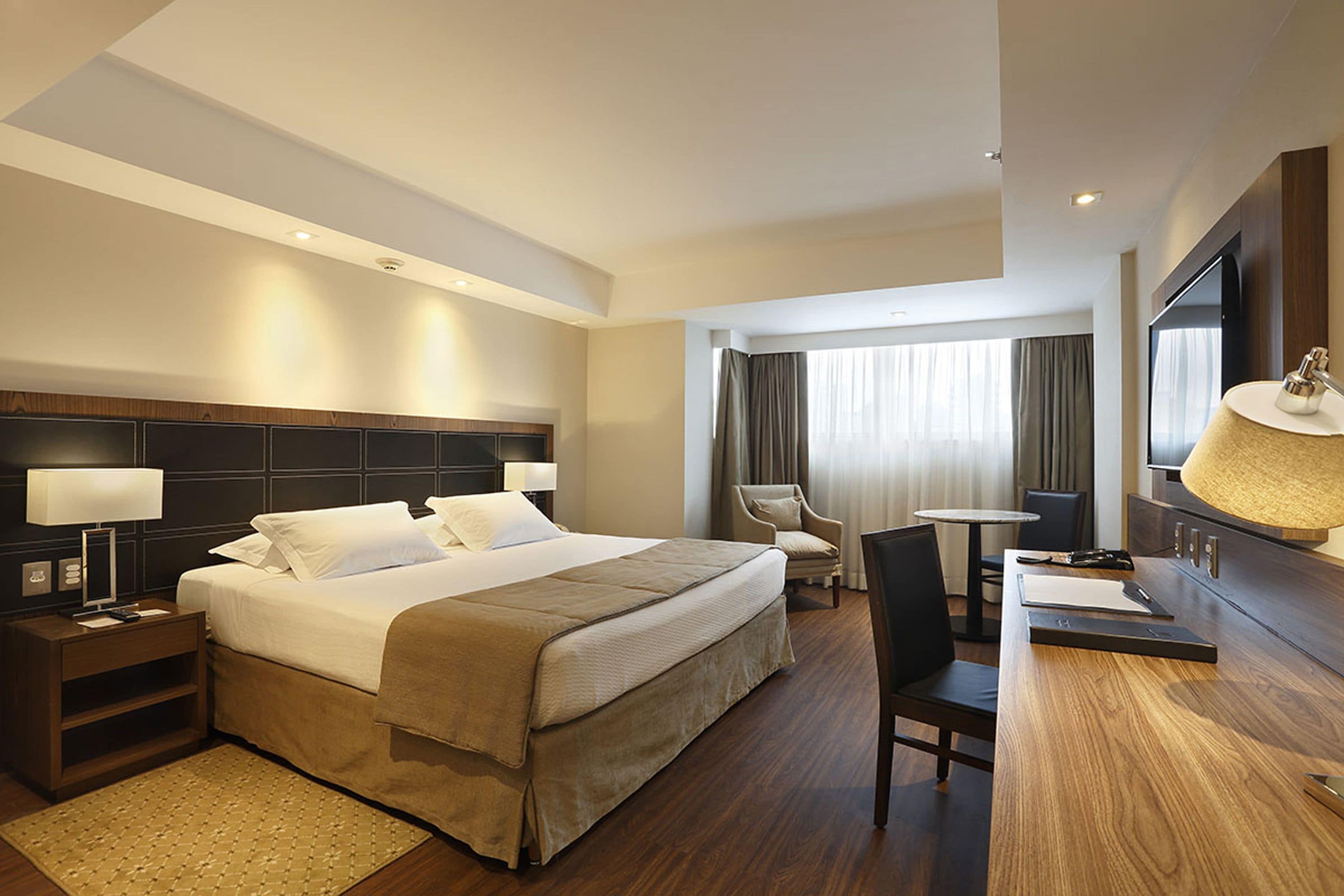 Habitación de hotel con una cama grande, un escritorio, una silla, un televisor y una ventana con cortinas.