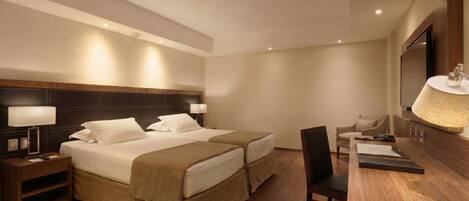 A hotel room with two beds, a desk, a chair, a TV, and a lamp.
